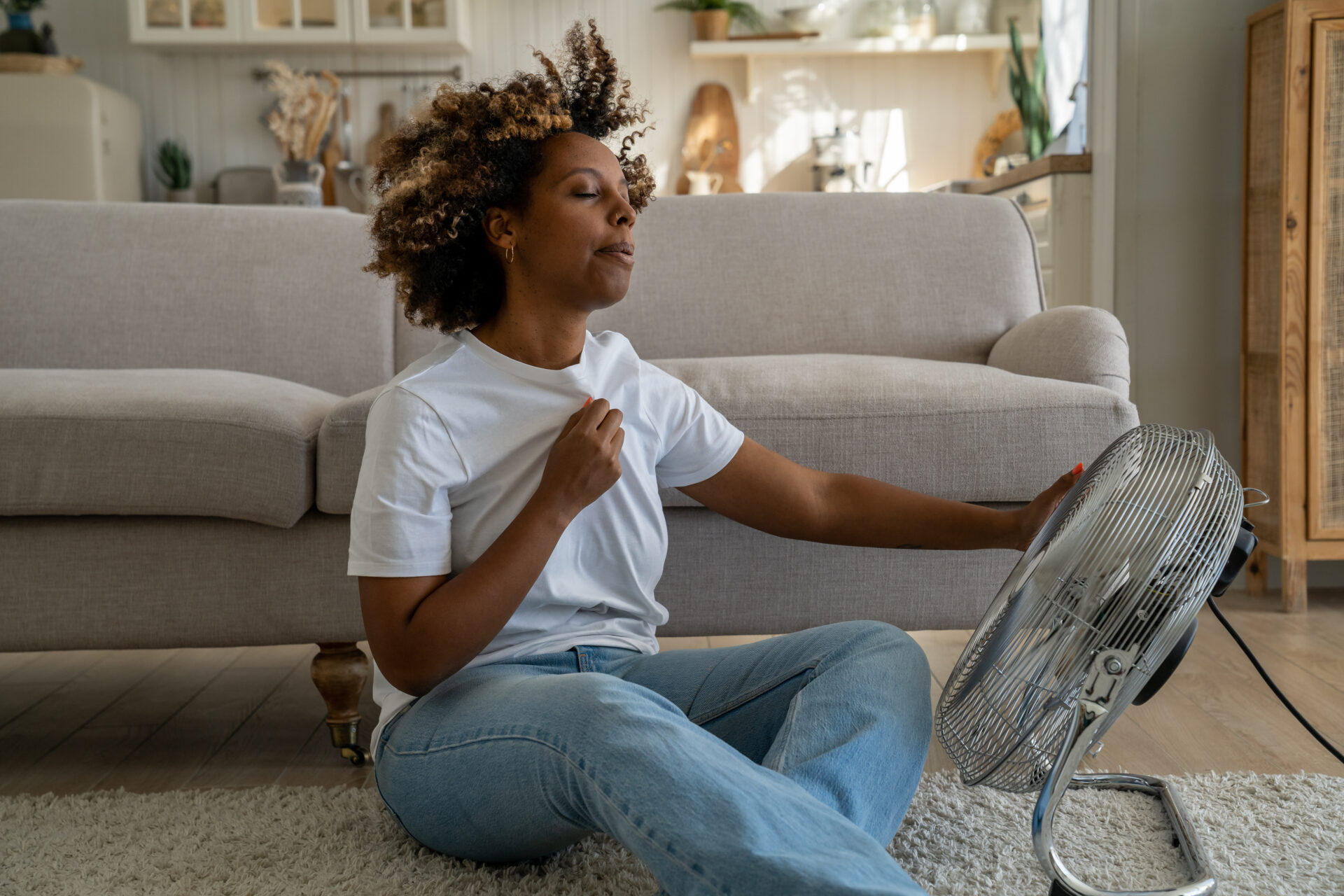 A dark-skinned woman in a white T-shirt cools down in front of a fan while waiting for the air conditioner repair