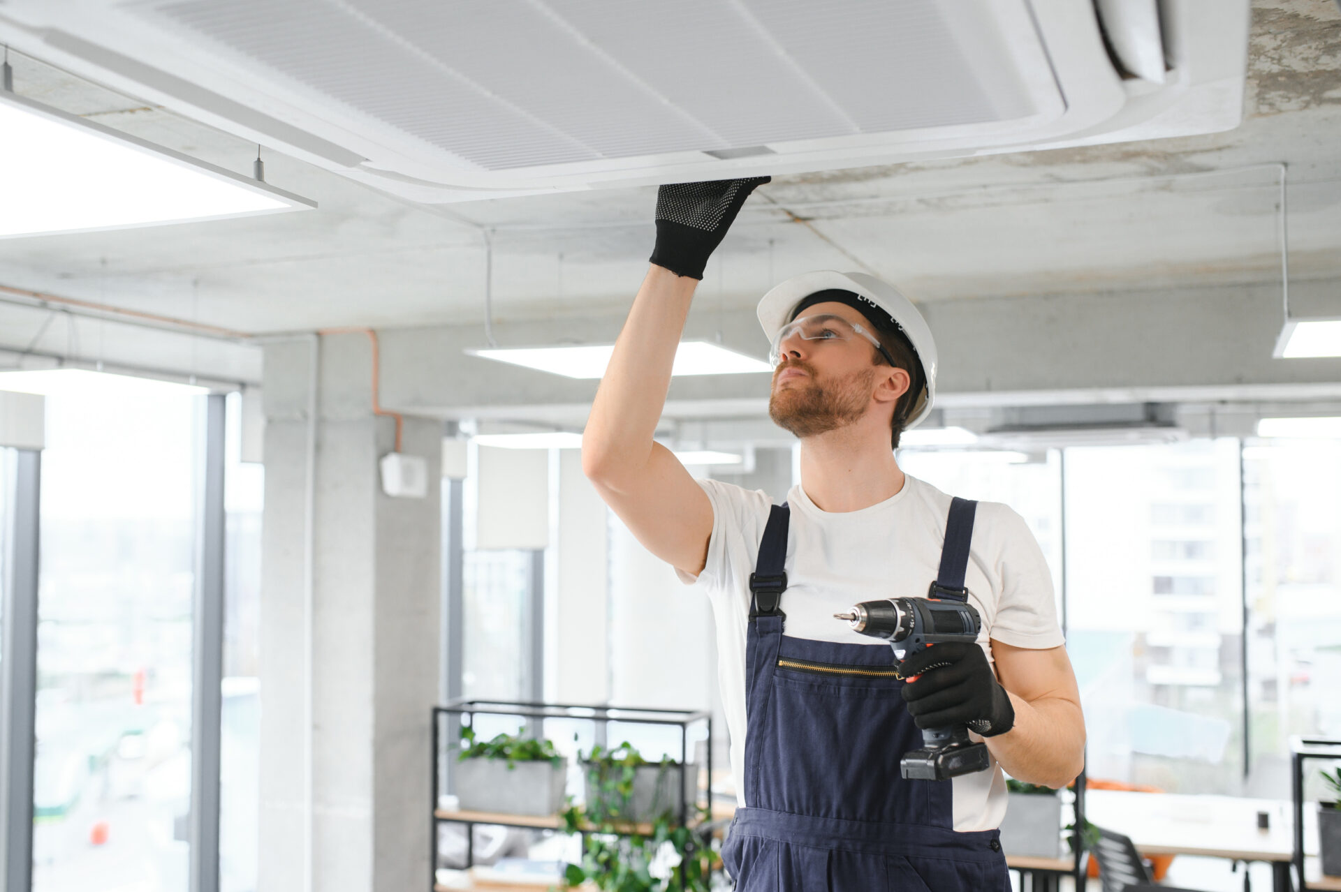 A worker in a blue uniform and white T-shirt with a drill checks an air conditioner repair