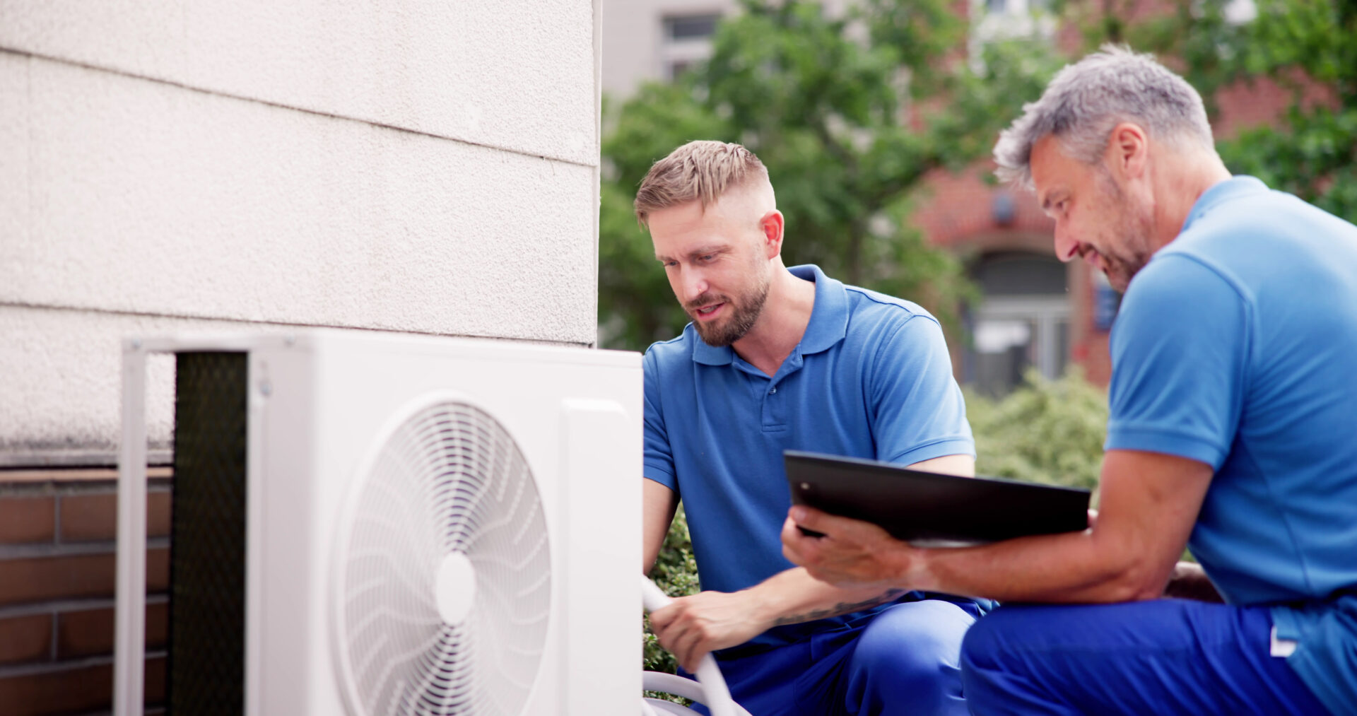 Two middle-aged men sit next to the wall in blue T-shirts and provide AC installation