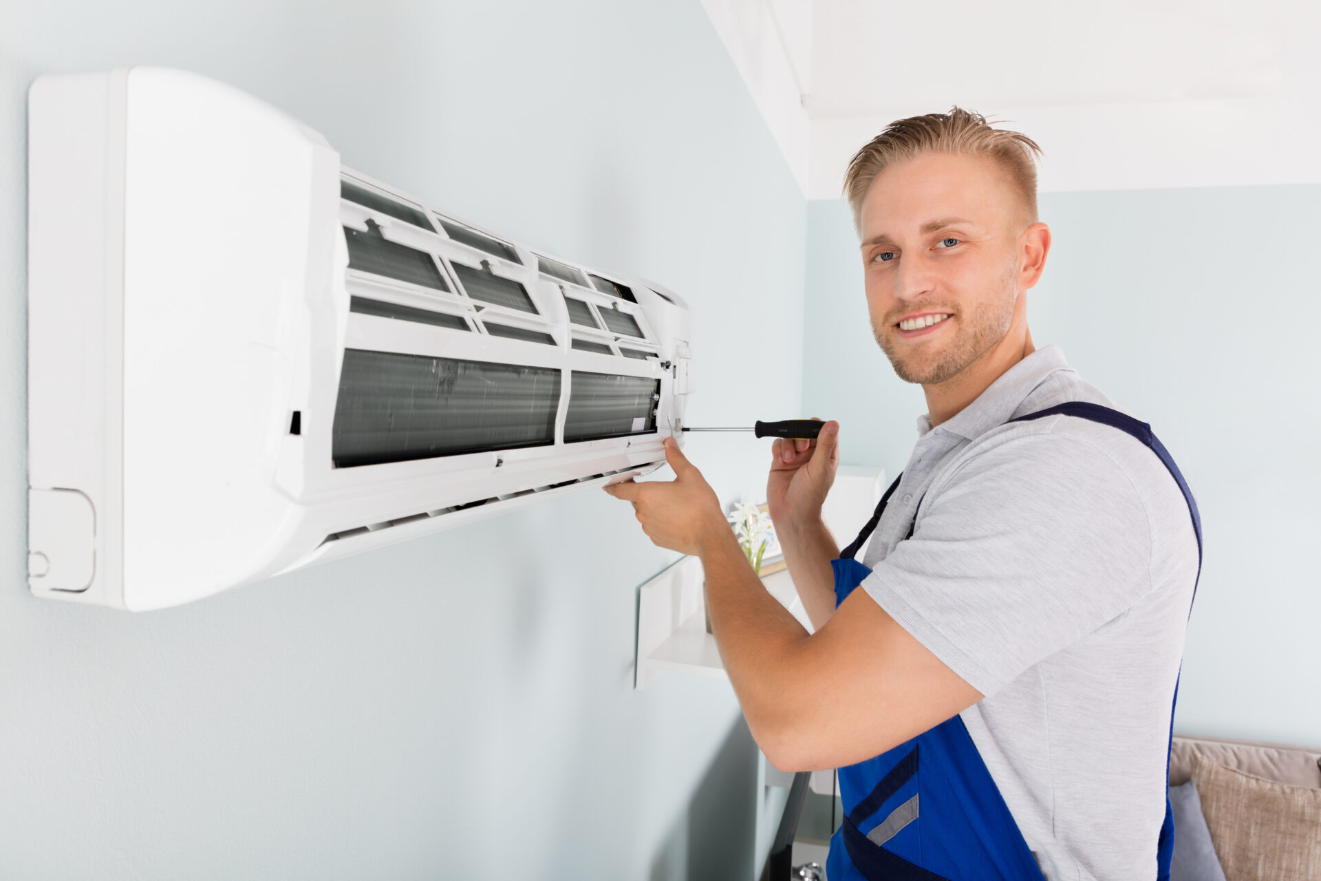 Good-looking male worker in uniform looking at camera and smiling while doing hvac repair with screwdriver